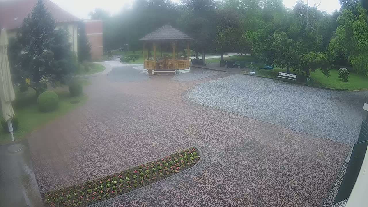 A rain-misted gravel courtyard features a wooden gazebo, landscaping, and a building in the background.