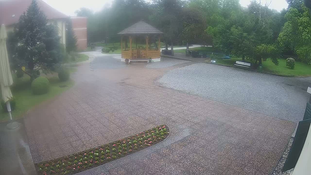 A gravel courtyard with a wooden gazebo, surrounded by green trees and bushes, in the rain.
