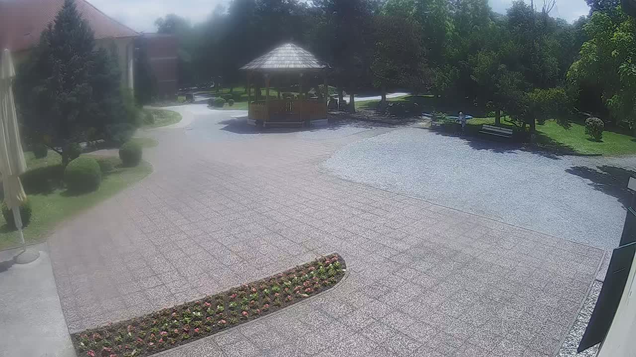 A paved courtyard with a wooden gazebo, flowerbeds, and a park-like setting in sunny weather.