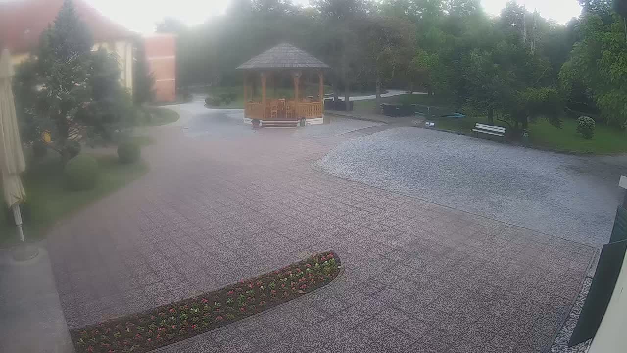A stone courtyard with a wooden gazebo and surrounding greenery is lightly covered in what appears to be hail under an overcast sky.