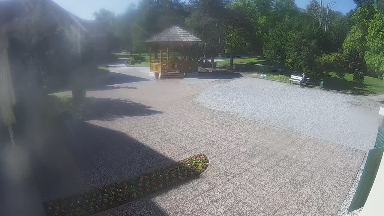 A paved courtyard with a wooden gazebo, a person sitting on a bench in a park, and flowerbeds under a sunny sky.