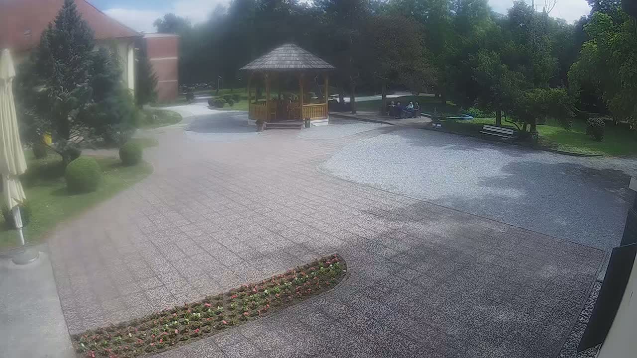 A paved courtyard with a wooden gazebo, flowerbeds, and park benches, surrounded by green trees and shrubs on a partly cloudy day.