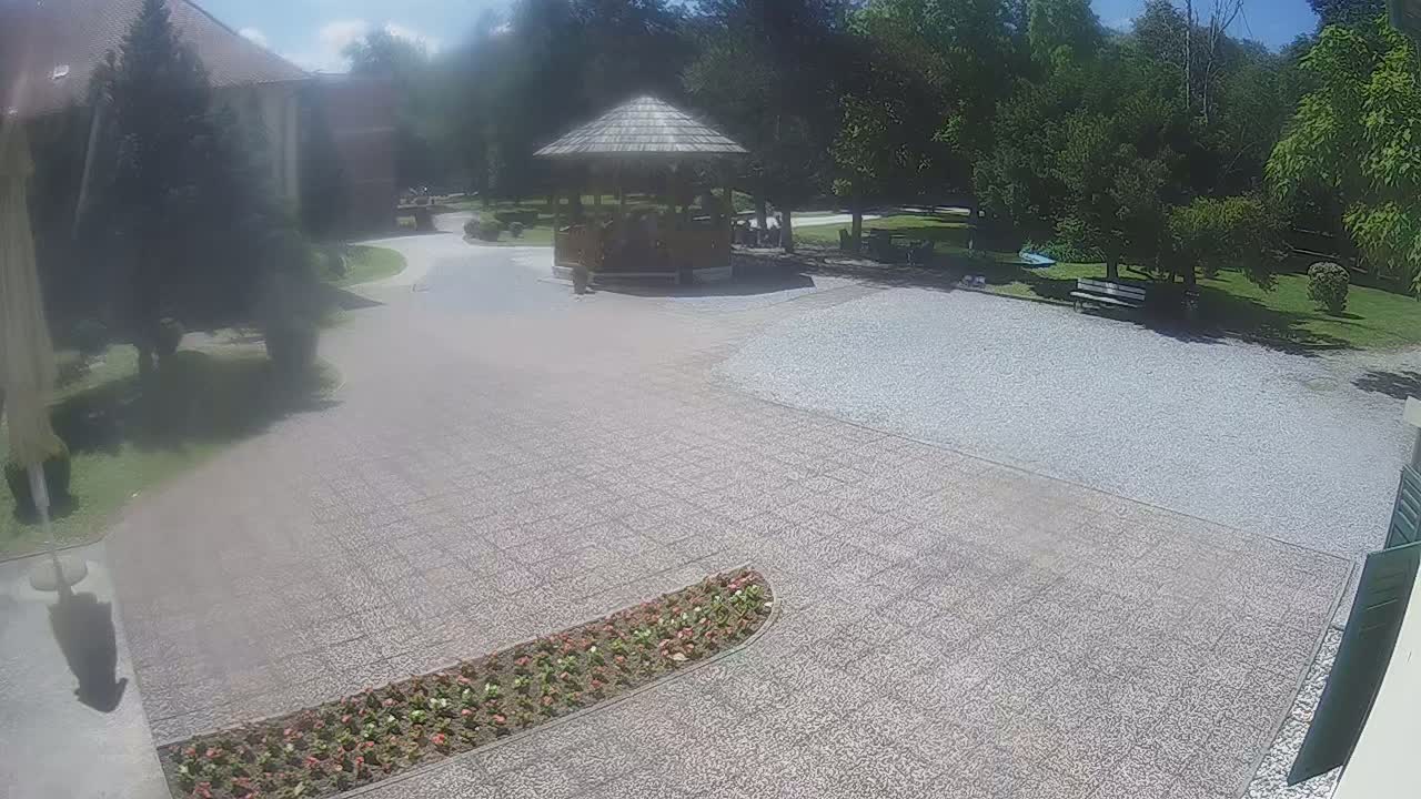 A paved courtyard with a flowerbed, a gazebo with people inside, a gravel area, and a park-like setting under a sunny sky.