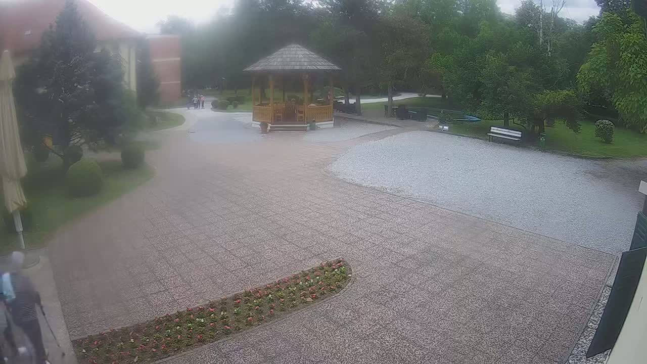 A hazy outdoor scene shows a paved courtyard with a wooden gazebo, a few benches, some landscaping, and a building in the background.