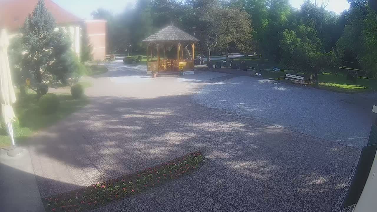 A gravel courtyard with a wooden gazebo, surrounded by trees and landscaping, on a sunny day.