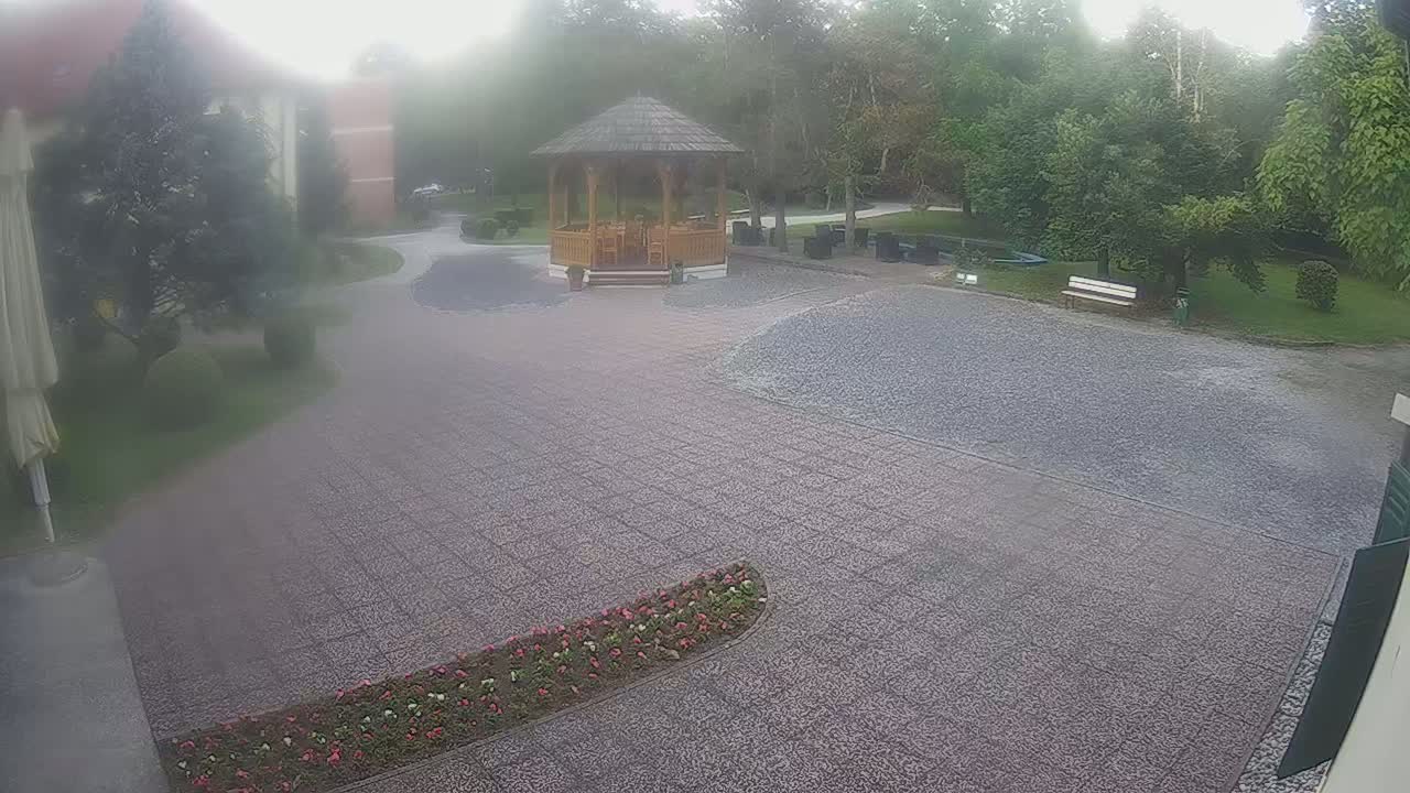 A misty outdoor scene shows a gravel courtyard with a wooden gazebo, landscaping, and a building partially visible in the background.