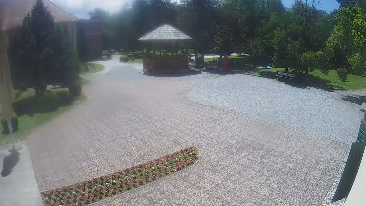 A gravel courtyard with a wooden gazebo and flowerbed is surrounded by green trees and a building under a partly sunny sky.