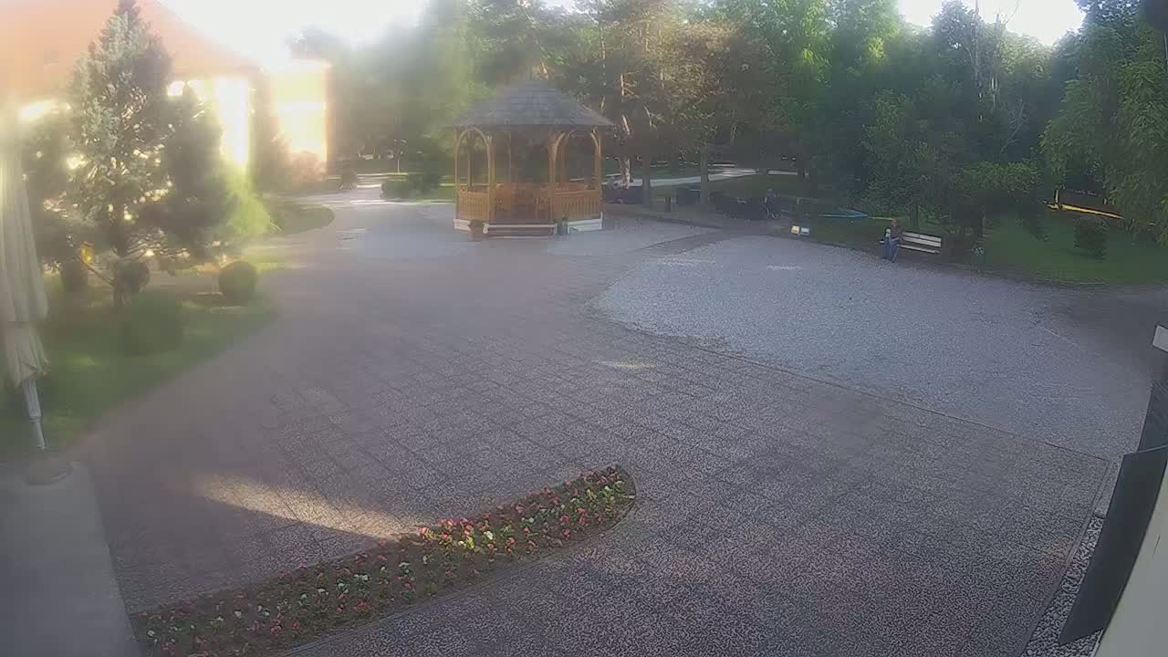 A hazy sunlit gravel courtyard features a wooden gazebo, landscaping, and a person sitting on a bench, surrounded by lush green trees.
