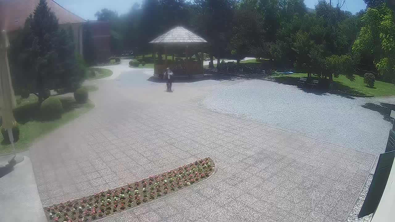 A person walks across a gravel courtyard toward a wooden gazebo on a sunny day, surrounded by lush green landscaping and trees.