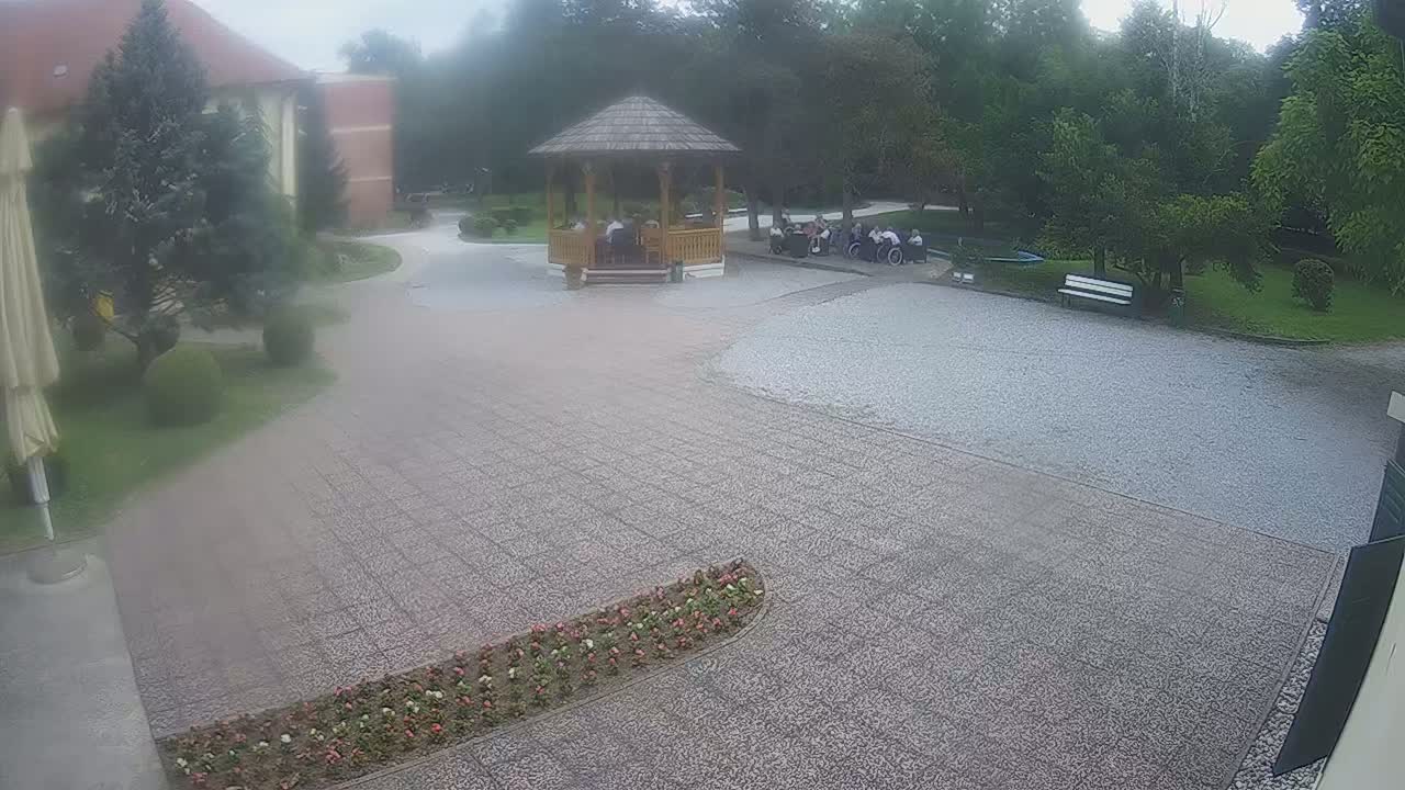 A hazy outdoor scene shows a gravel courtyard with a wooden gazebo containing several people, surrounded by a building, green lawns, and flowerbeds.