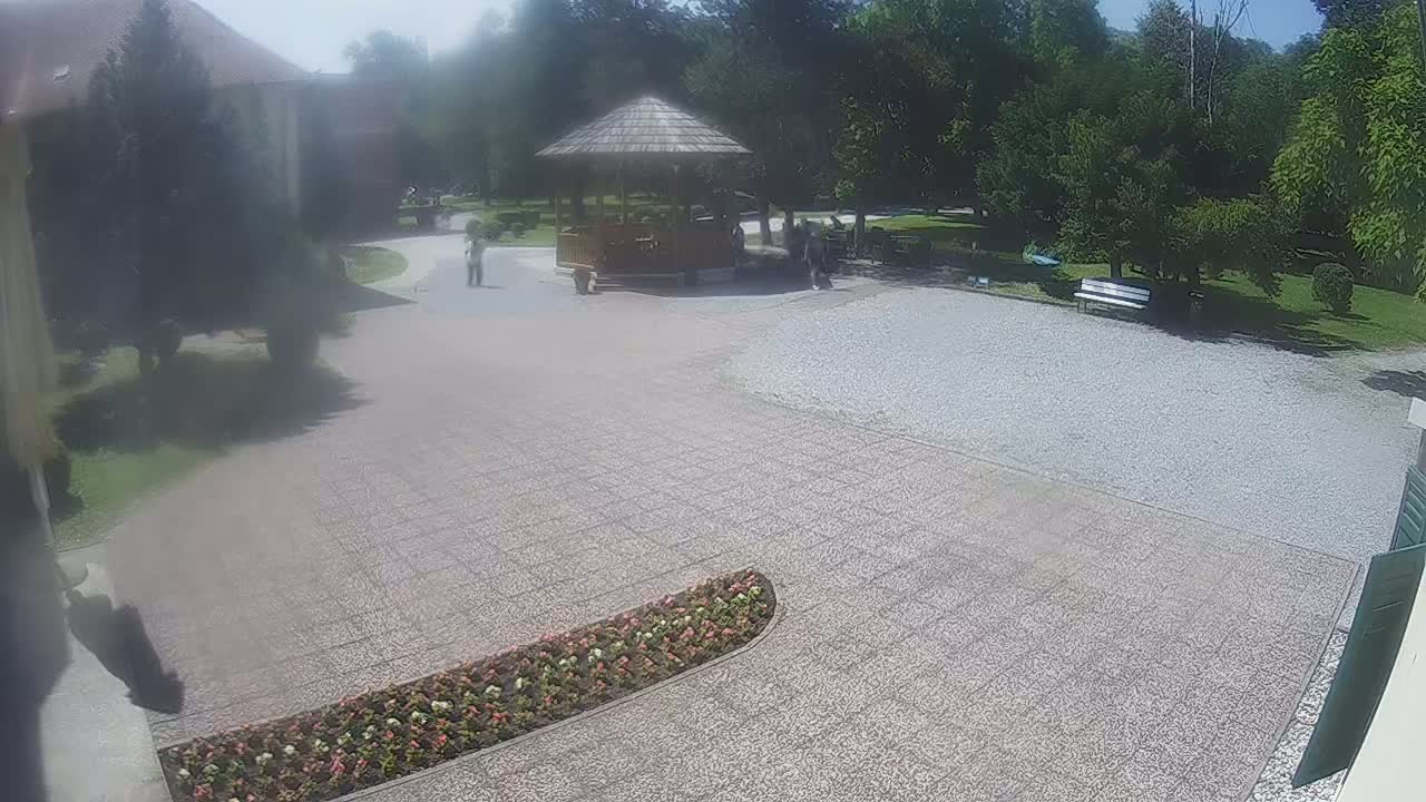 A gravel courtyard with a wooden gazebo and flowerbeds, surrounded by trees on a sunny day.