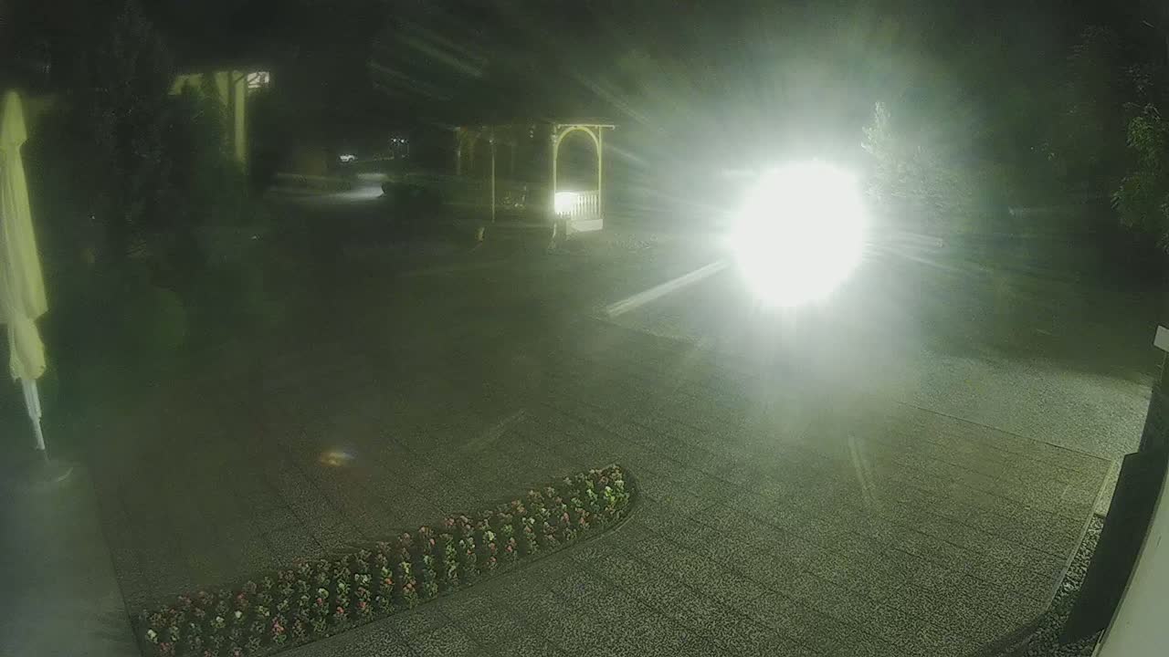 A nighttime, brightly lit gravel area with a gazebo and flower bed is visible, with a bright light source obscuring much of the view.