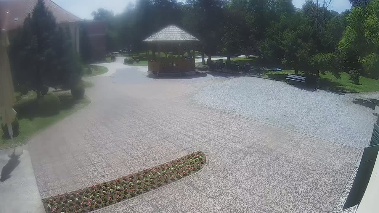 A gravel courtyard with a wooden gazebo, flowerbeds, and a building in the background on a sunny day.