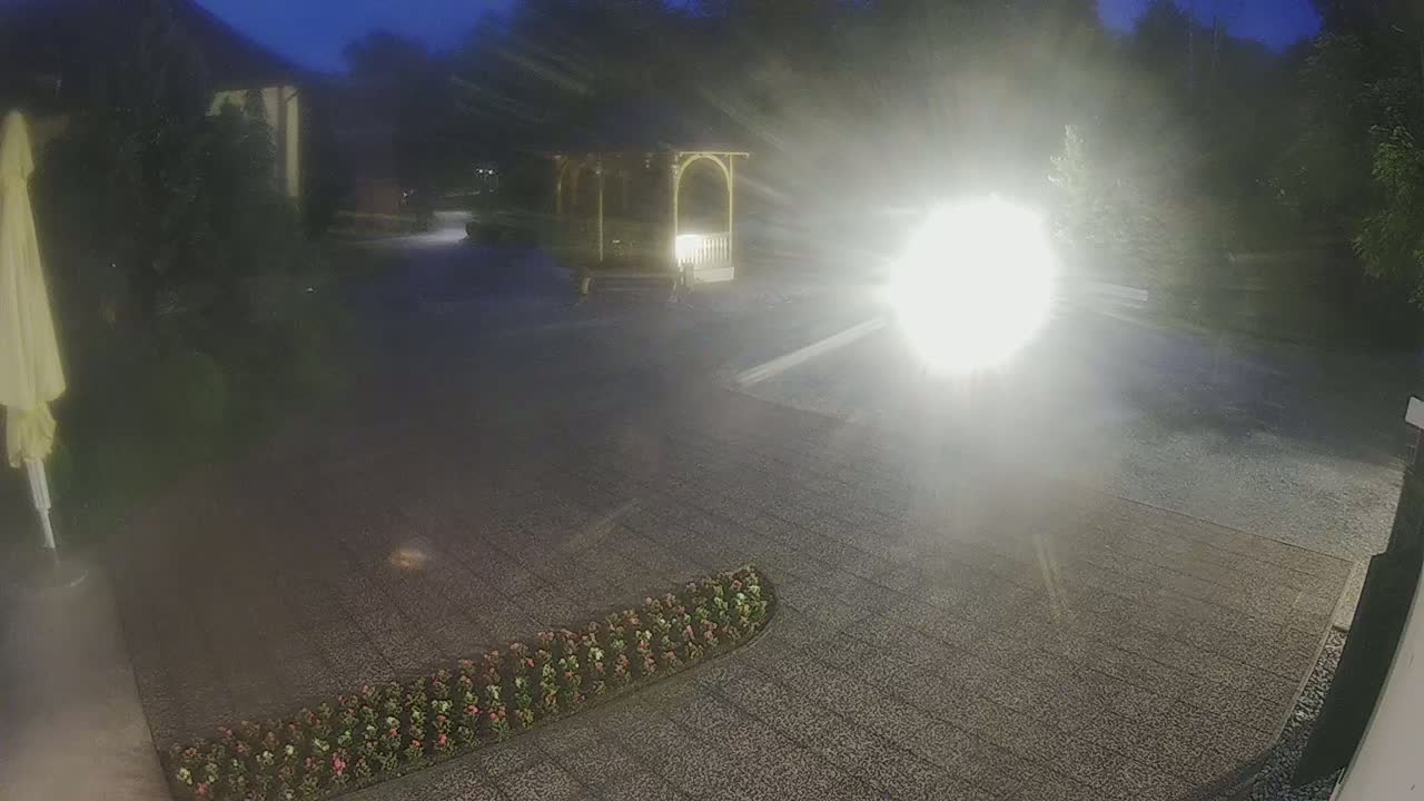 A nighttime, brightly lit gravel area shows a gazebo and a flower bed, with a bright light source obscuring much of the center of the image.