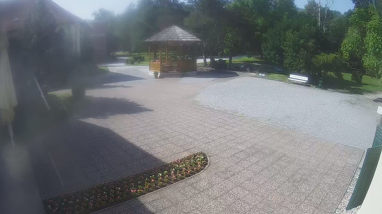 A paved courtyard with a wooden gazebo, a flower bed, a gravel area, and a bench, all under a sunny sky.