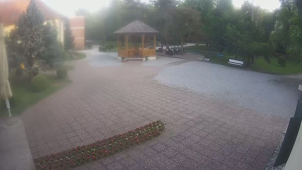 A hazy view shows a gravel courtyard with a wooden gazebo and several people sitting inside, surrounded by manicured lawns and trees.