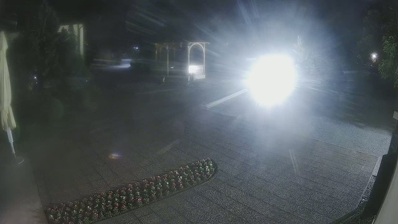 A nighttime, brightly lit outdoor scene shows a paved area with a flower bed, a gazebo, and a very bright light source in the middle distance.