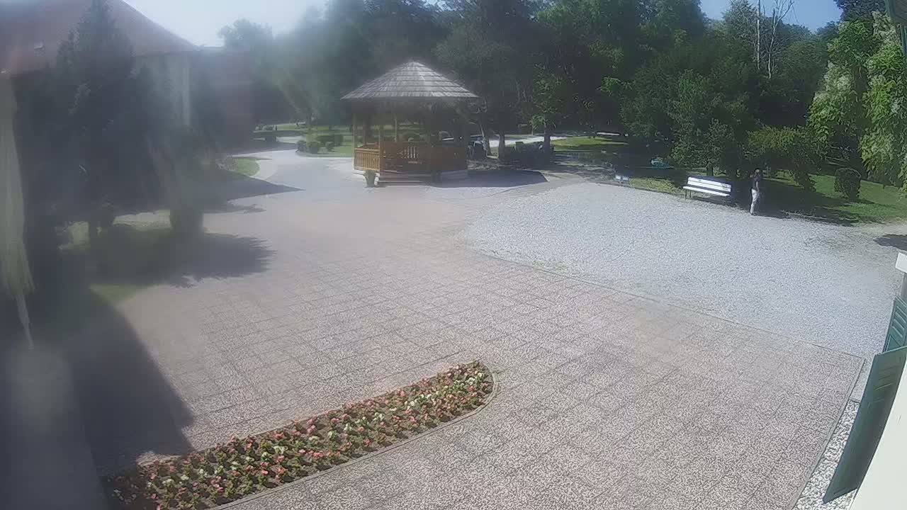 A gravel courtyard, partially paved, features a wooden gazebo, a flower bed, a park bench, and a person walking near some trees on a sunny day.