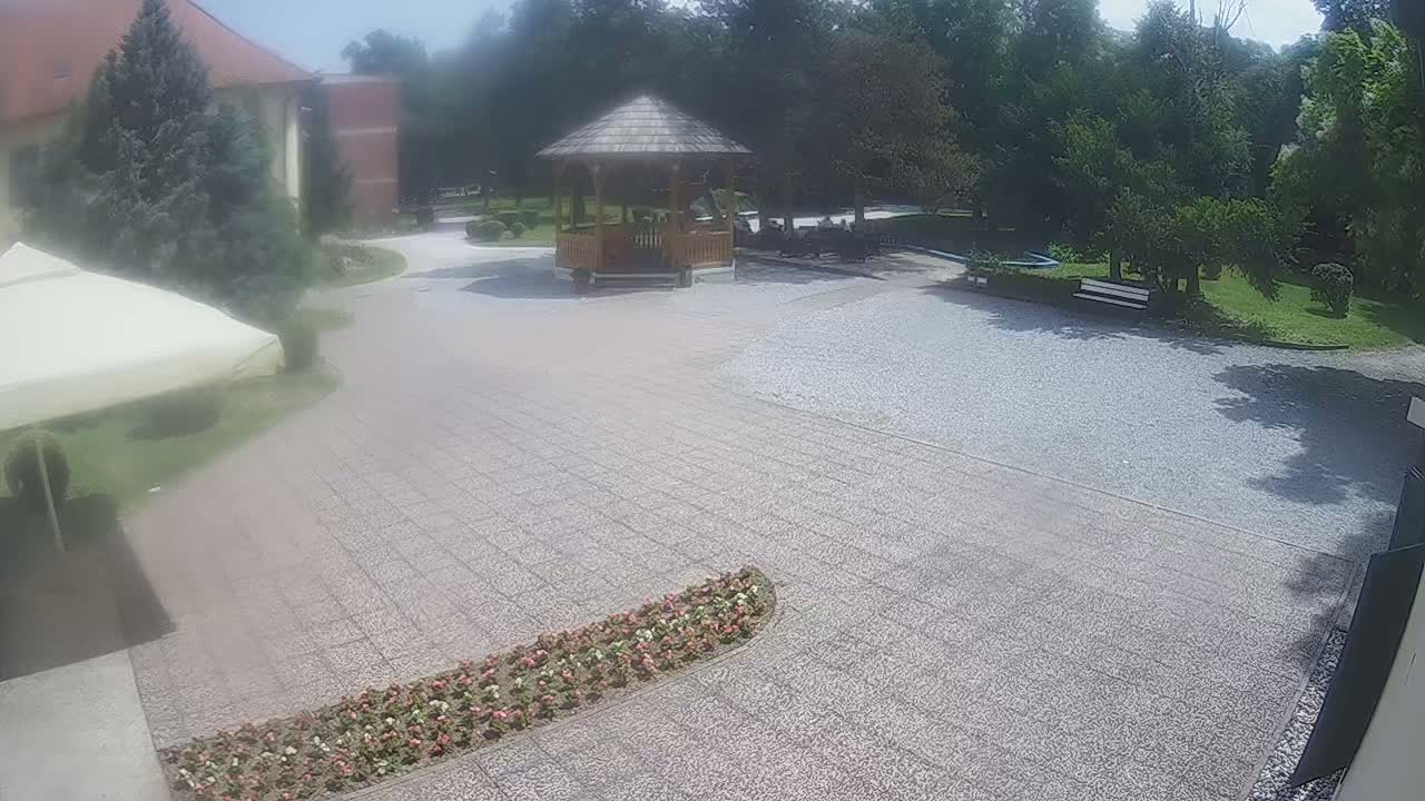 A gravel courtyard with a wooden gazebo, surrounded by trees and flowerbeds, on a sunny day.