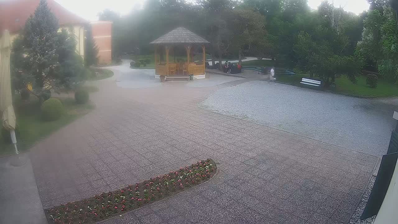 A hazy outdoor scene shows a gravel courtyard with a wooden gazebo, a building in the background, and several people sitting near the gazebo.