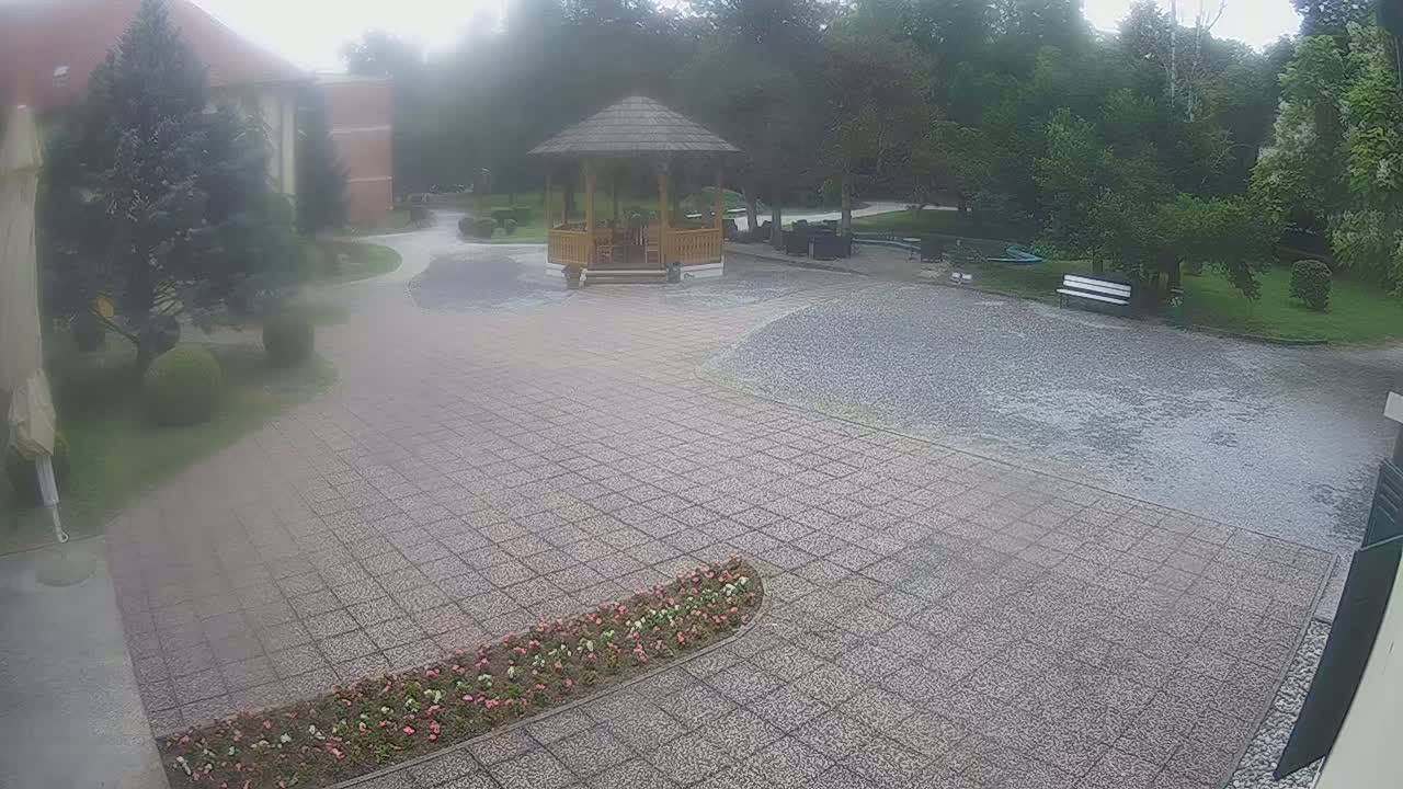 A paved courtyard with a wooden gazebo, surrounded by a park with lush green trees and shrubs on a foggy day.
