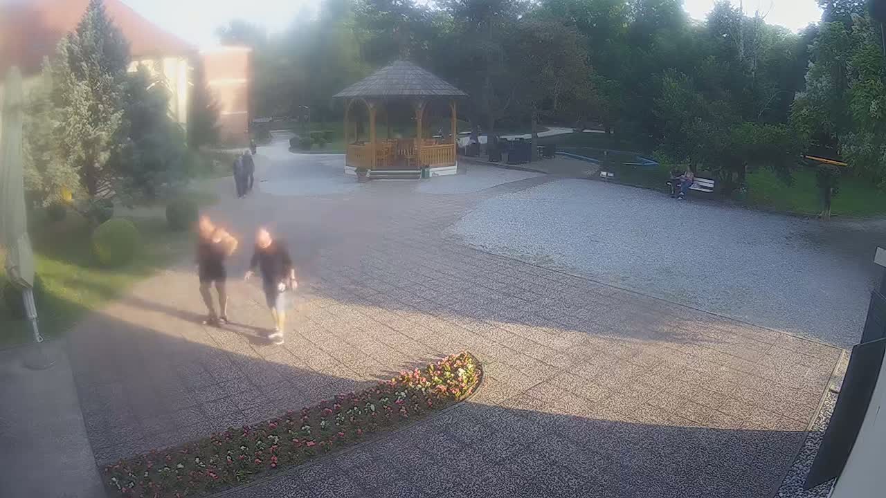 A hazy, sunlit outdoor scene shows two people walking toward the camera in a courtyard with a gazebo, some shrubs, and a few other people in the distance.