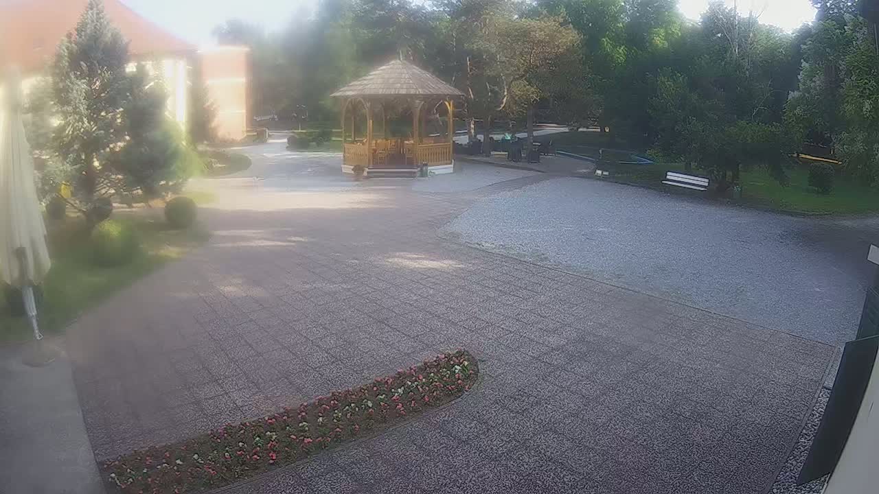 A gravel courtyard with a wooden gazebo, surrounded by trees and flowerbeds, on a hazy, sunny day.