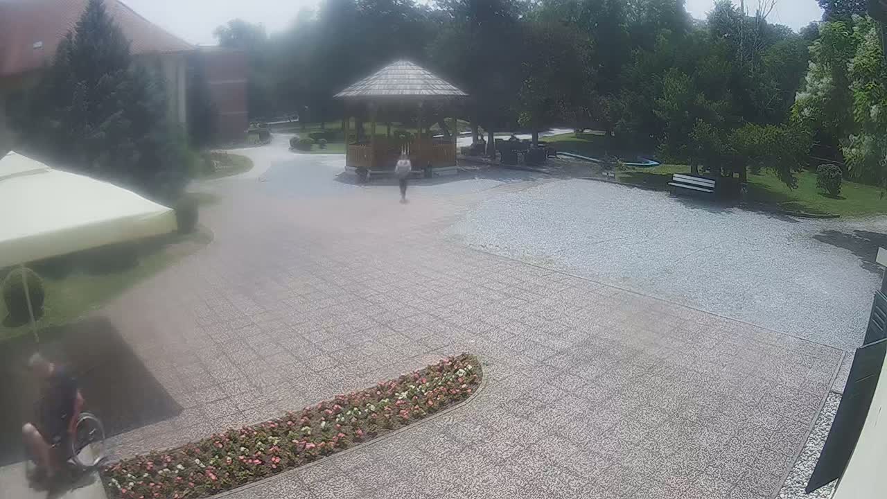A person in a wheelchair and another person are walking near a gazebo in a gravel courtyard on a hazy day.