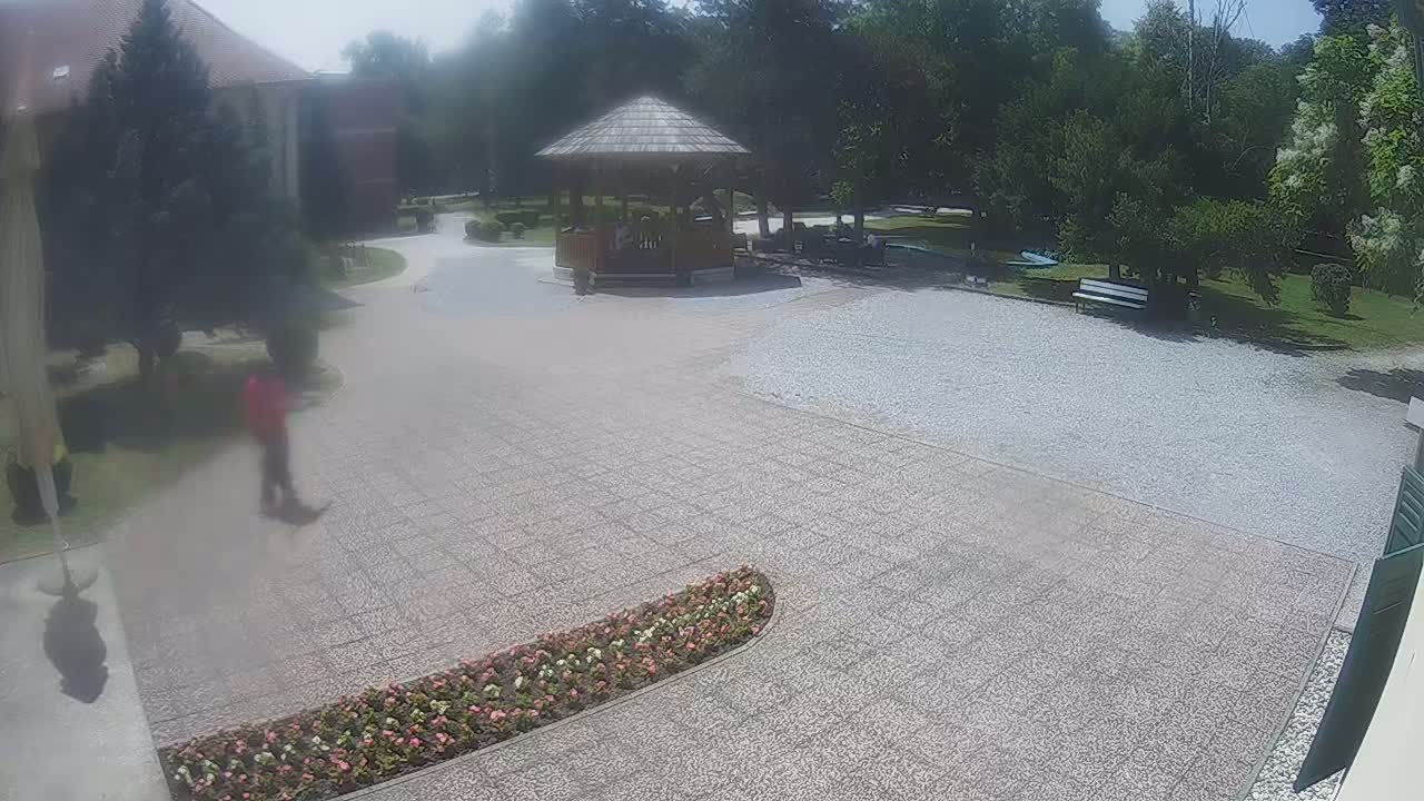 A paved courtyard with a wooden gazebo, a few people, landscaping, and a building in the background on a sunny day.