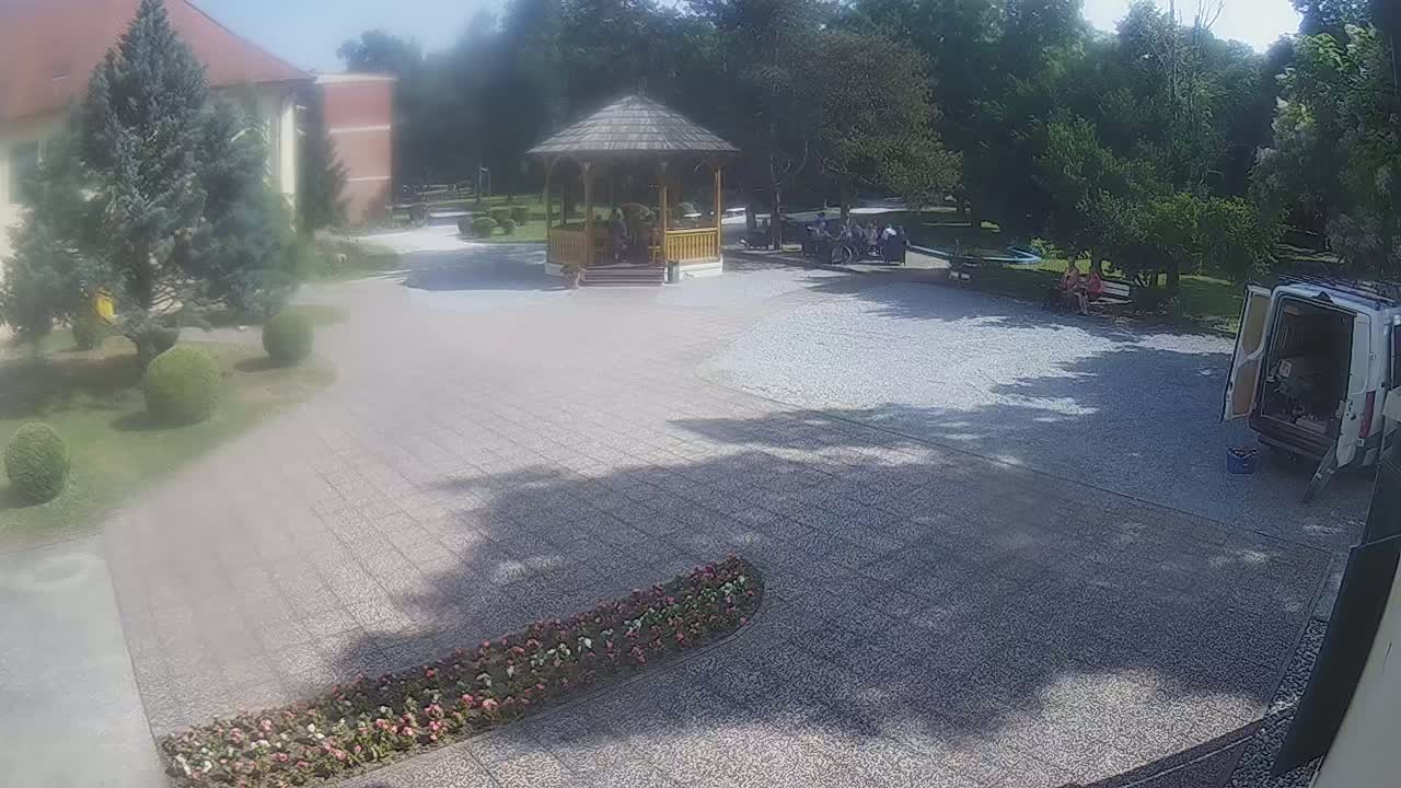 A gravel courtyard with a wooden gazebo and several people, surrounded by green trees and bushes under a sunny sky, features a van parked nearby and flowerbeds lining the path.
