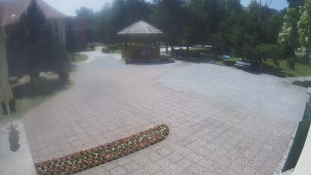 A paved courtyard with a gazebo and flowerbed, surrounded by trees and a building, on a sunny day.