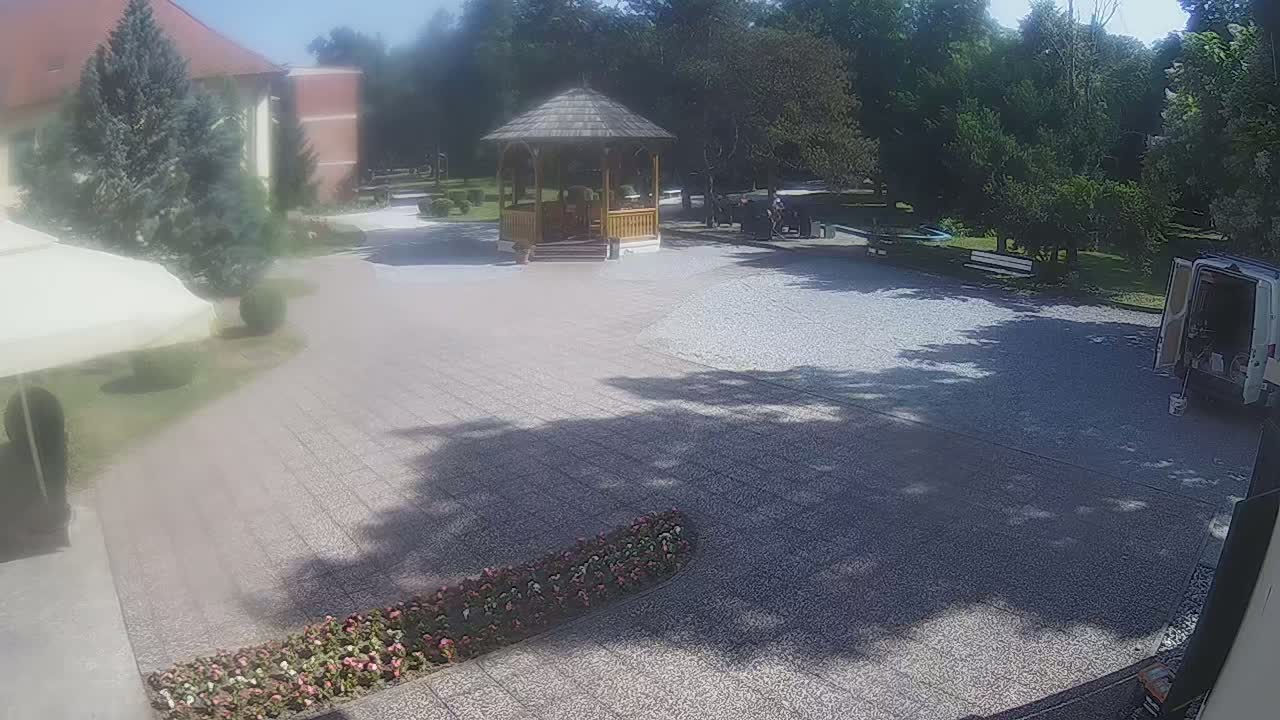 A gravel courtyard with a wooden gazebo, surrounded by trees and shrubs, on a sunny day.
