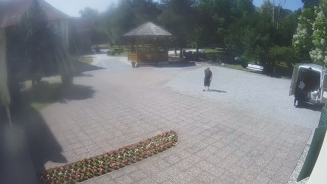 A sunny day reveals a gravel courtyard with a wooden gazebo, a person walking, a van with its door open, and a flowerbed.