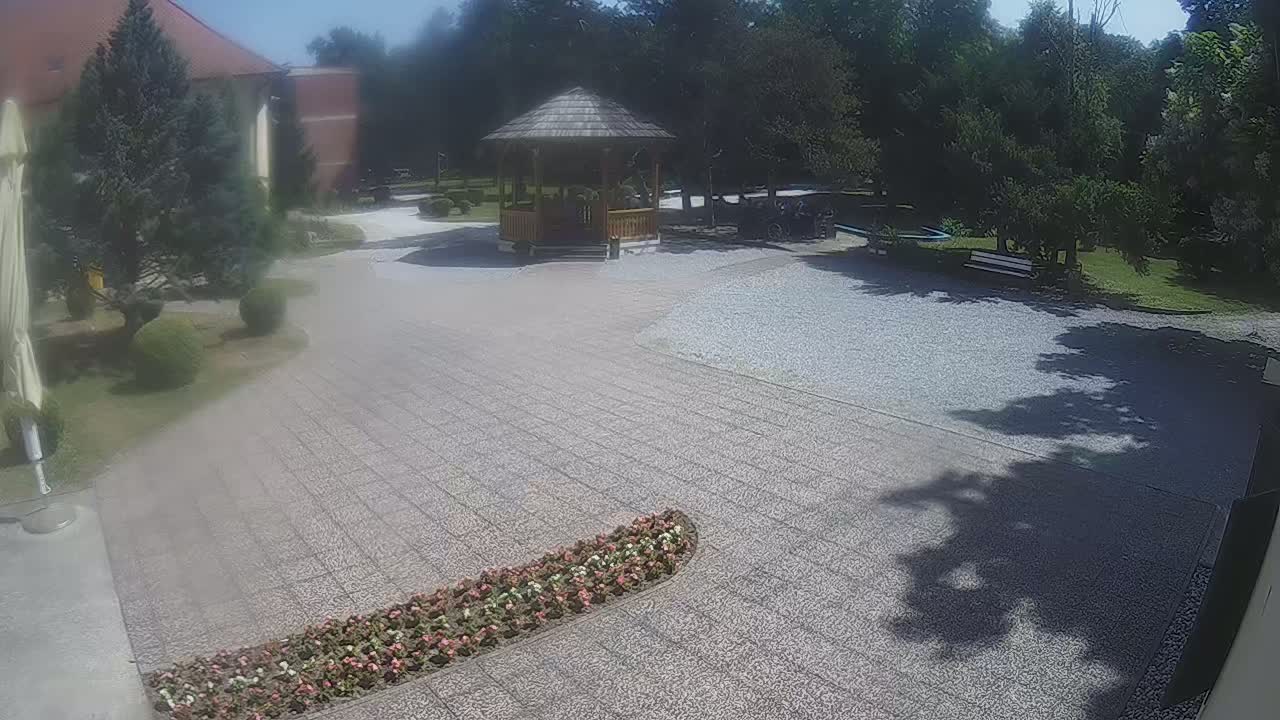 A gravel courtyard with a wooden gazebo, flowerbeds, and trees, on a sunny day.