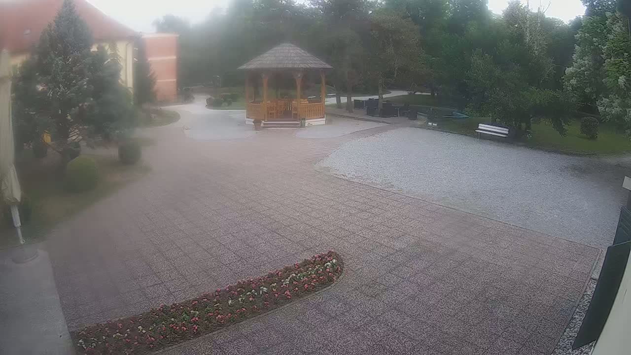A gravel courtyard with a wooden gazebo, flowerbeds, and trees, on a sunny day.