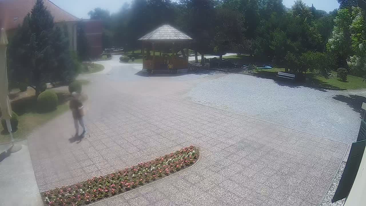 A person walks across a paved courtyard on a sunny day near a wooden gazebo and some buildings, with surrounding landscaping including flowers and trees.
