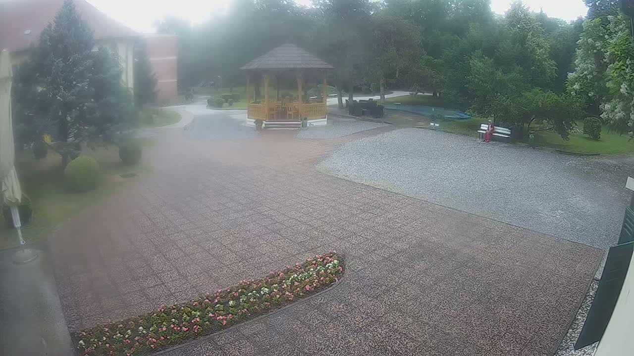 A hazy view of a gravel courtyard with a wooden gazebo, surrounded by greenery and a person sitting on a bench, in misty weather.