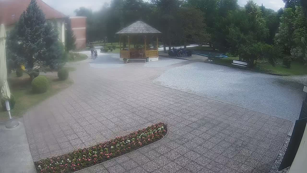 A paved courtyard with a wooden gazebo, some people, and landscaping is visible under an overcast sky.