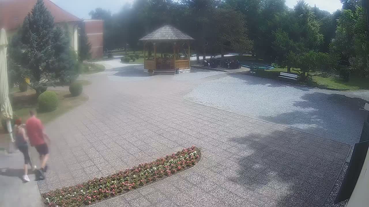 A couple walks across a paved courtyard toward a wooden gazebo in a park-like setting on a partly sunny day.