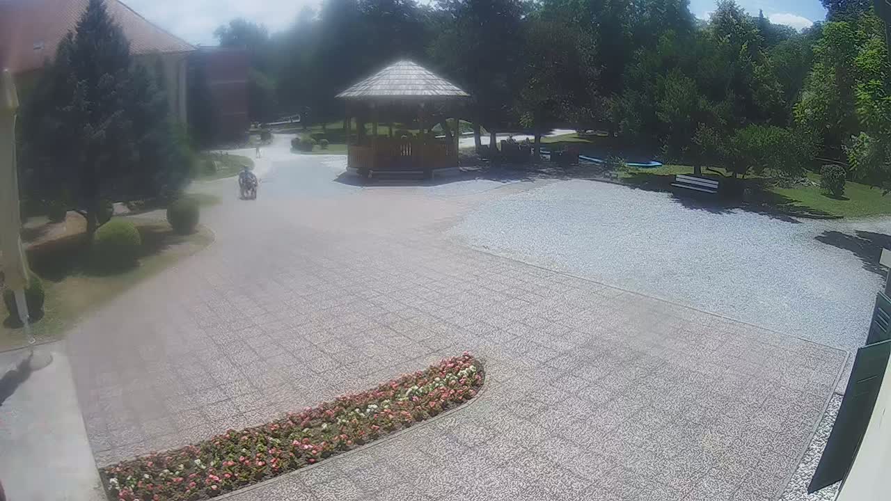 A person walks on a gravel path toward a wooden gazebo in a sunny courtyard with lush green trees and flowerbeds.
