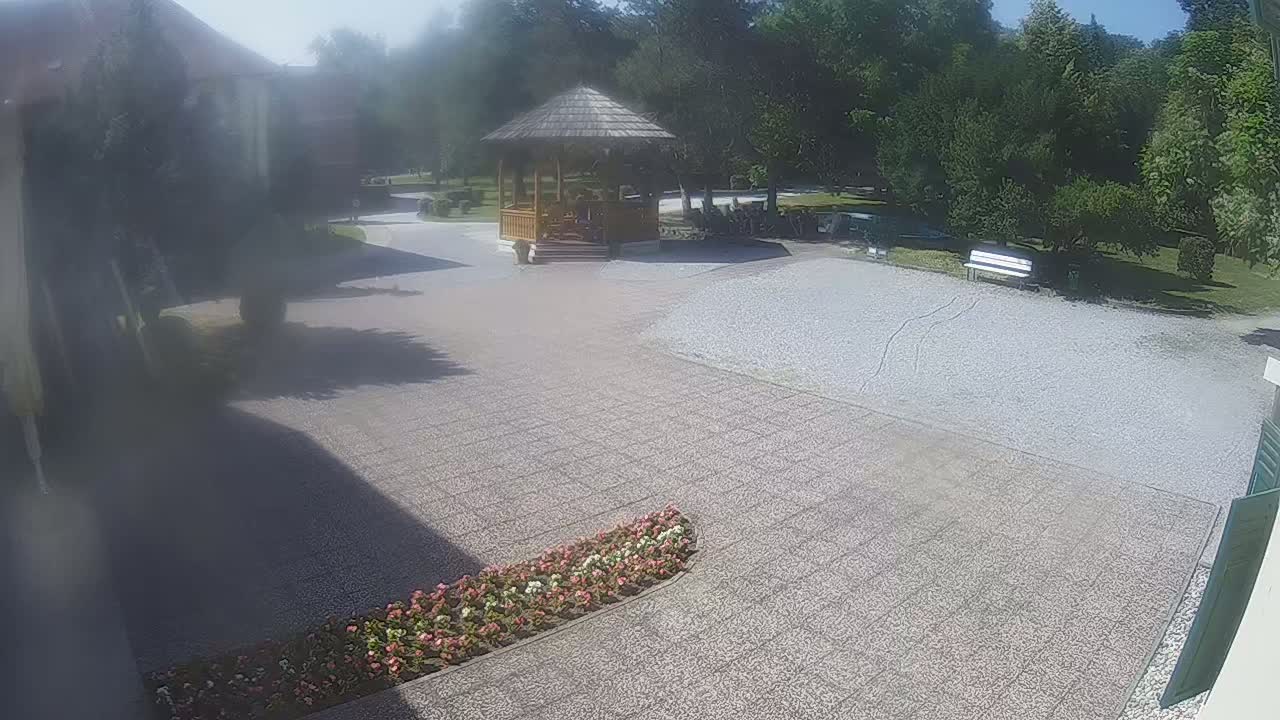 A gravel courtyard with a wooden gazebo, a bench, and flowerbeds, surrounded by trees on a sunny day.