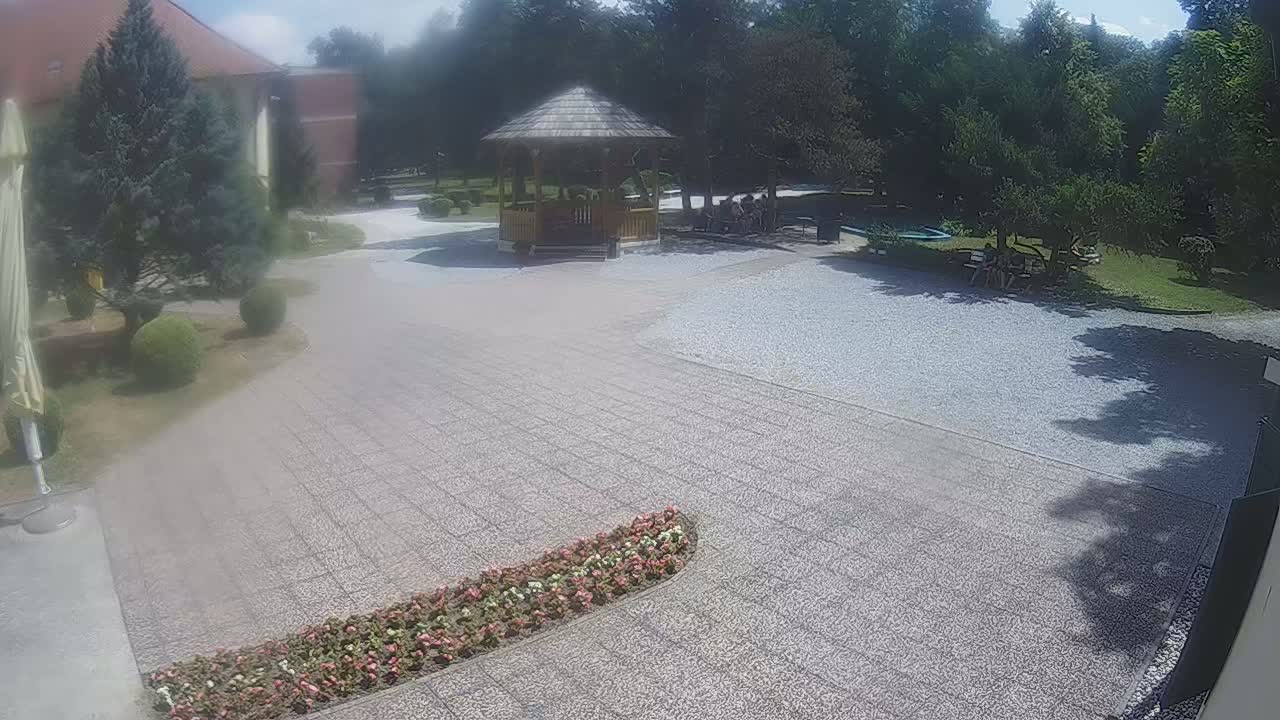 A gravel courtyard with a wooden gazebo and several people sitting beneath trees on a sunny day.