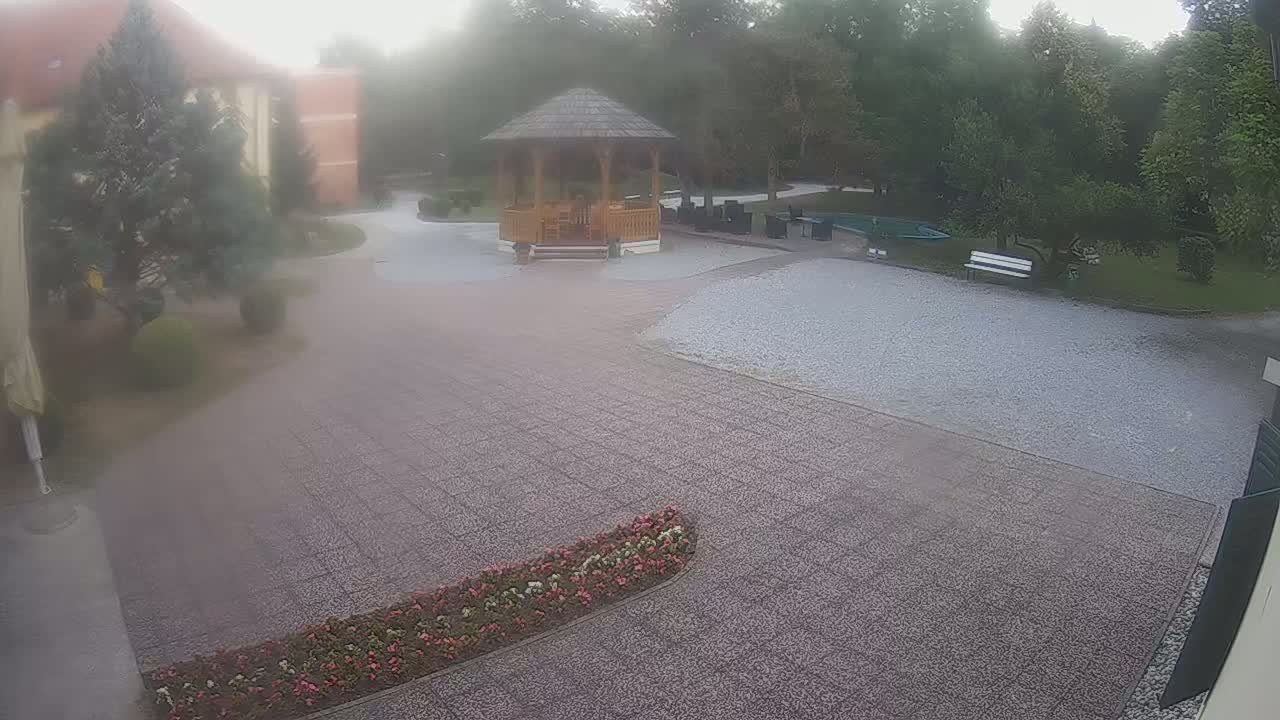 A hazy view shows a courtyard with a wooden gazebo, a gravel area, flowerbeds, and trees under an overcast sky.