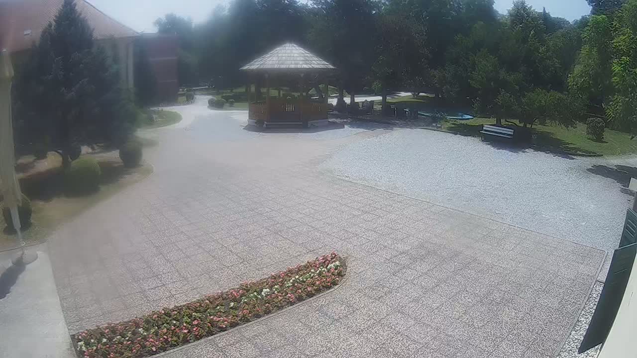 A gravel courtyard with a wooden gazebo, flowerbeds, and trees, on a sunny day.