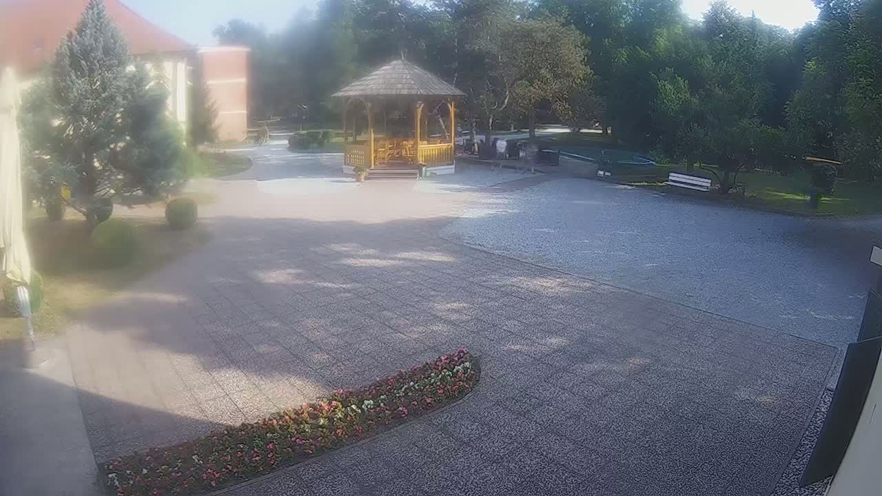 A gravel courtyard with a wooden gazebo, surrounded by trees and flowerbeds, on a partly sunny day.