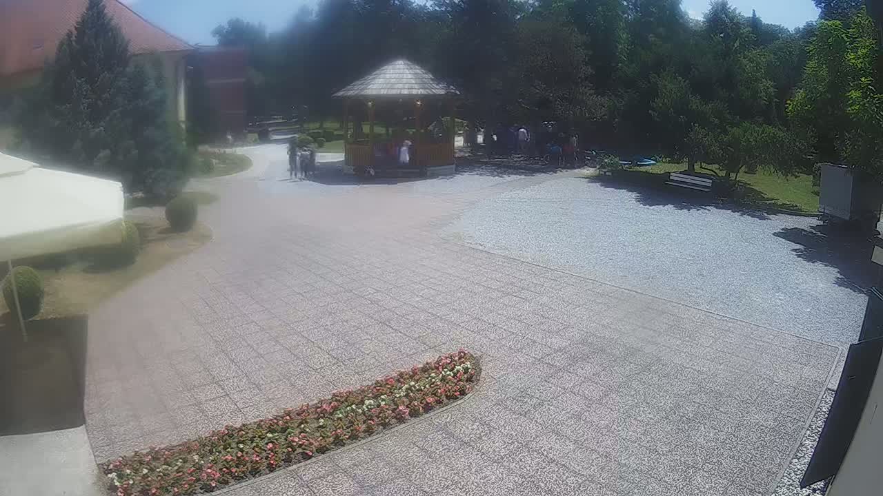 A gravel courtyard with a wooden gazebo and several people, under a sunny sky, is surrounded by greenery and a building.