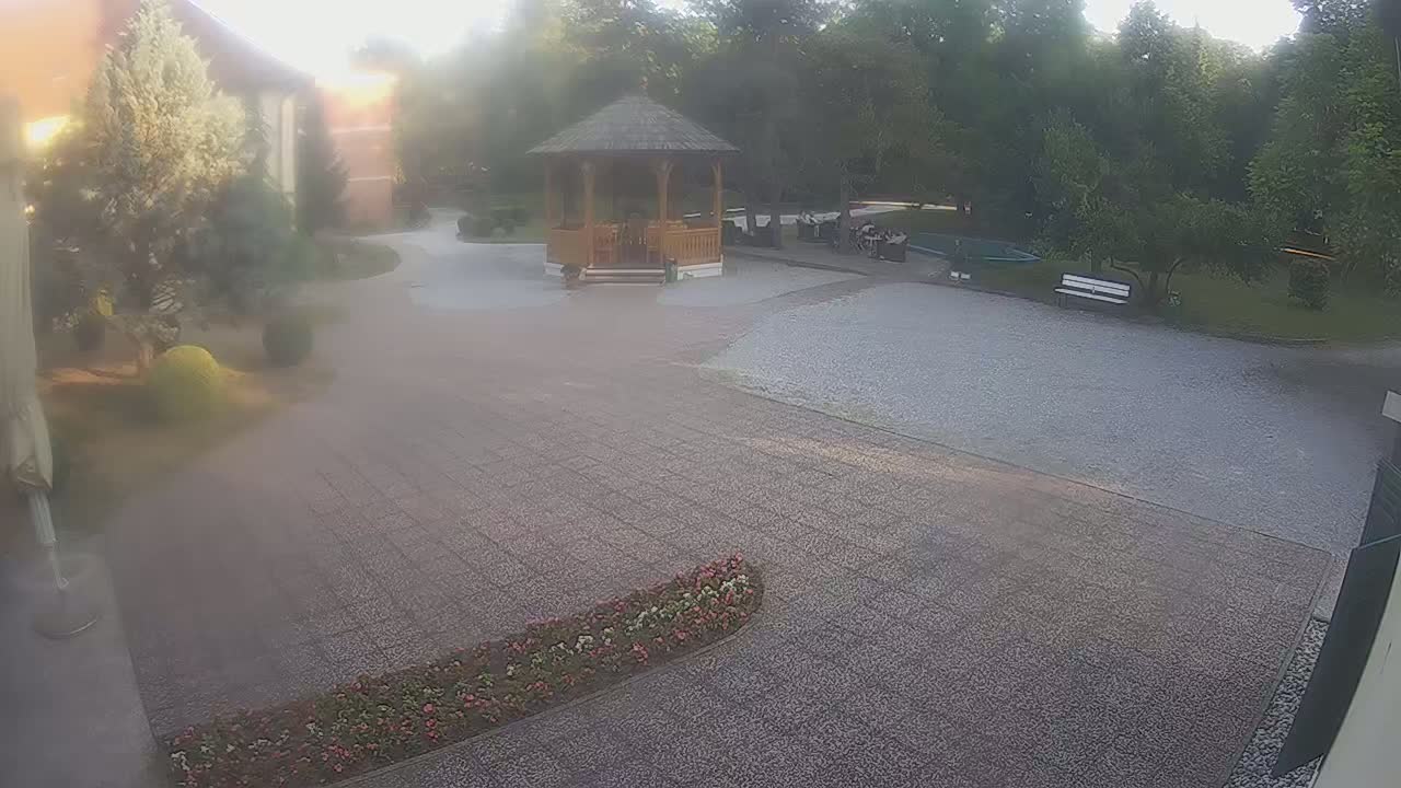 A hazy, outdoor scene shows a gravel courtyard with a wooden gazebo, a few benches, landscaping, and a building in the background.