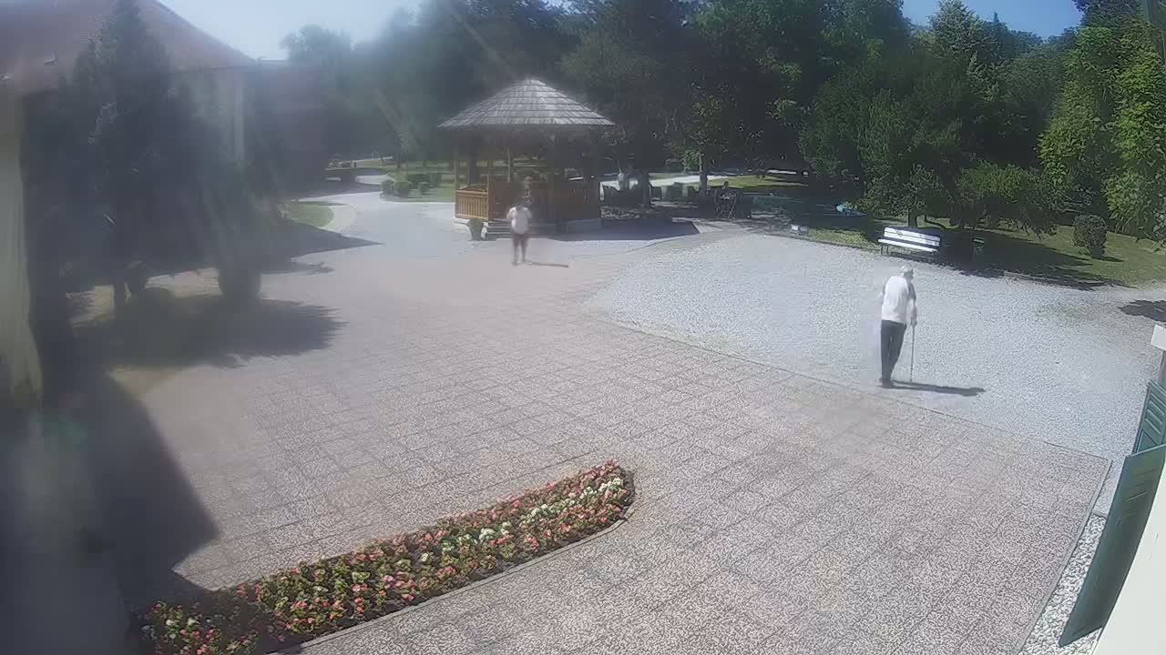 A sunny day shows three people walking on a gravel courtyard near a gazebo and some trees, with a flowerbed visible in the foreground.