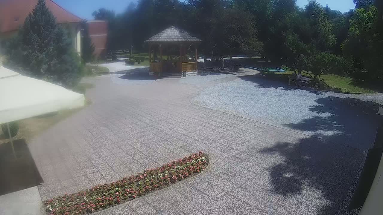 A gravel courtyard with a wooden gazebo, flowerbeds, and a partially visible building under a sunny sky.