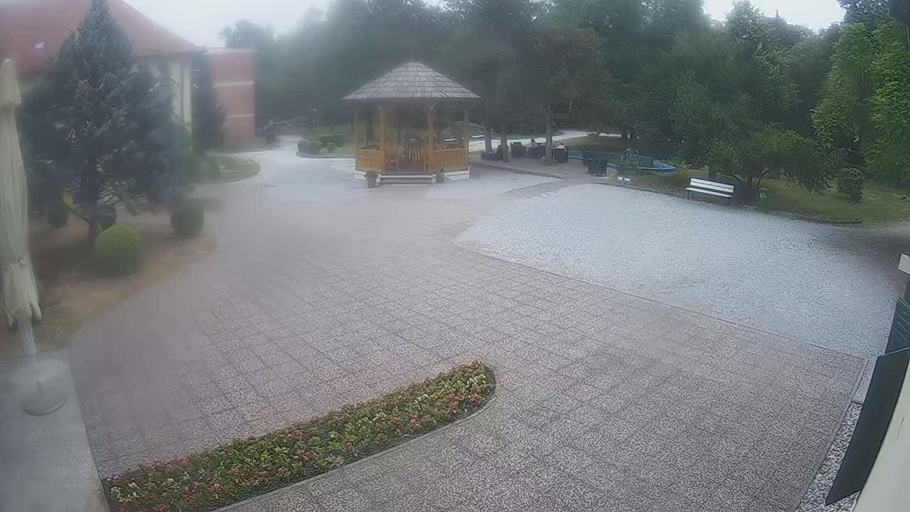 A gravel courtyard with a wooden gazebo, surrounded by trees and landscaping, on a foggy day.